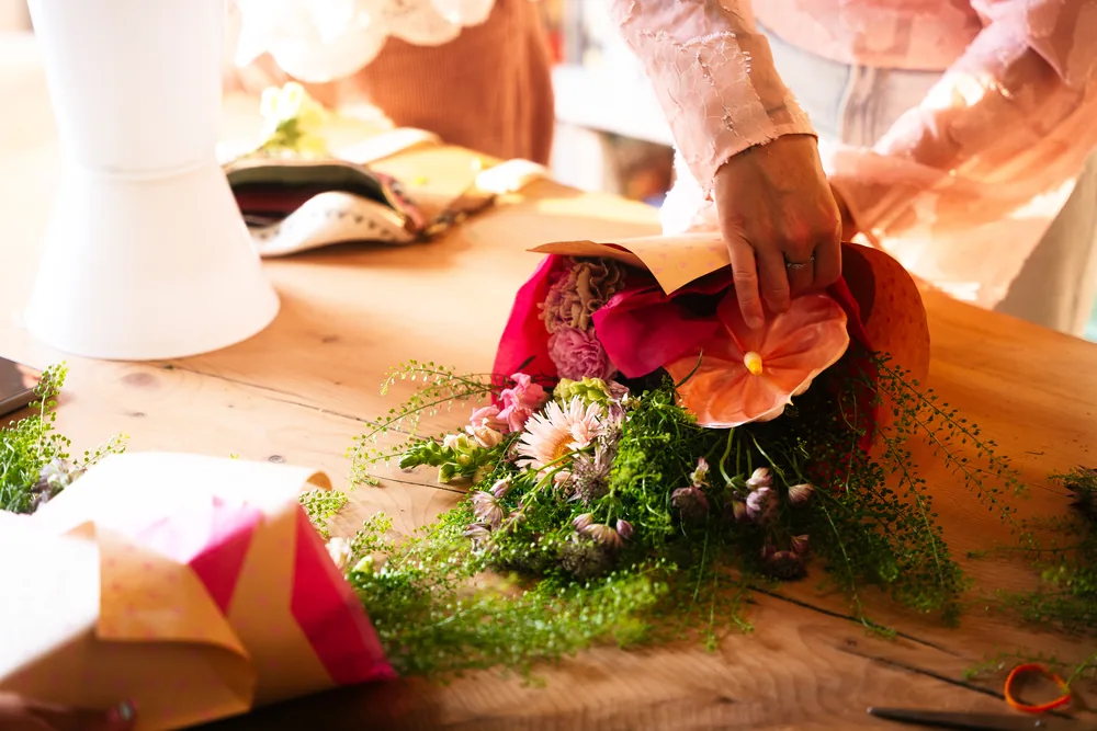 Hands arranging a bouquet of pink and orange flowers on a sunlit wooden table.