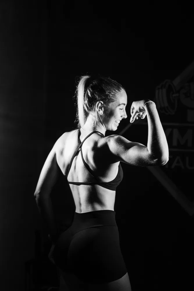 Hannah flexing a bicep and laughing, shot from behind in a dark gym, lit in high contrast black and white.