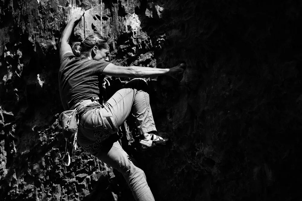 A climber in black and white, high on the limestone wall at Gordale Scar, reaching for a hold with one leg stretched out across the rock.